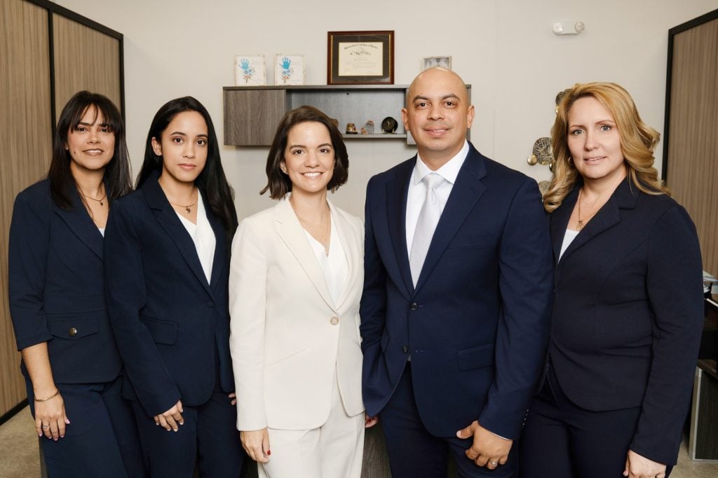 Five people wearing business suits stand together in an office, looking at the camera. Shelves, a framed certificate, and office furniture are visible in the background.