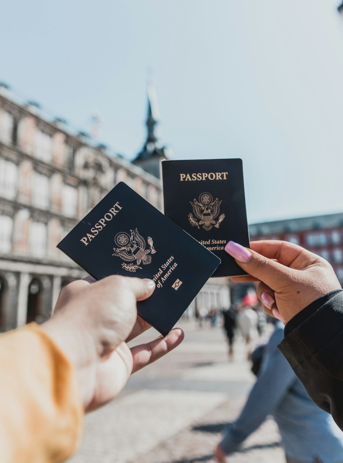 Two people hold up United States passports outdoors in a city square, with a historic building and spire visible in the background.